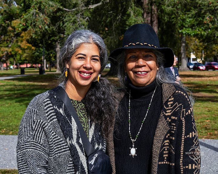 Two women smiling outdoors. One in a hat and patterned coat; the other in a patterned shirt and sweater. Trees in background.