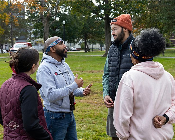 Four people conversing outdoors on a grassy area; one gestures, others listen.