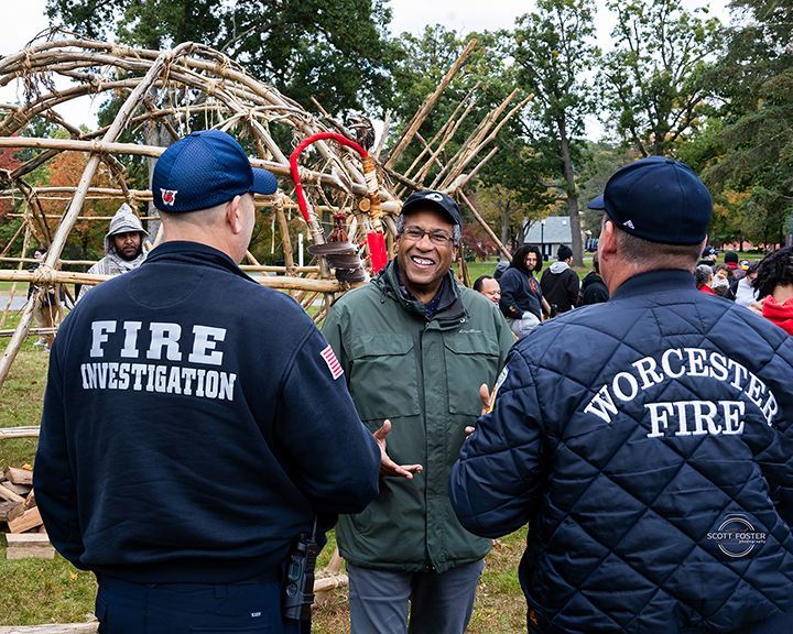 Two firefighters in dark jackets converse with a man in a green jacket outdoors. A wooden structure is in the background.