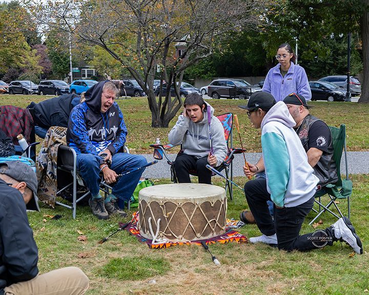 Group of people playing a large drum outdoors. Some are singing, others listening, fall day.