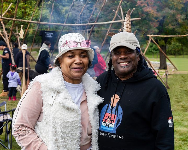 Two people smiling in front of a wooden structure, possibly an outdoor event.