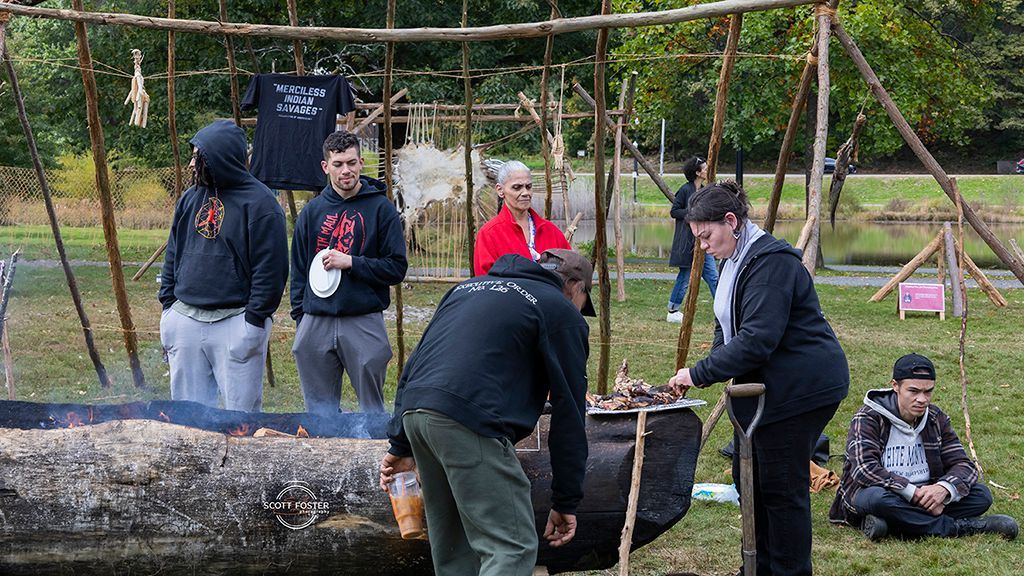 People tending a fire outdoors, near a wooden structure. Some look on as a woman adds to the fire.