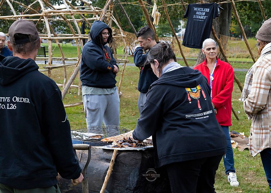 People preparing food outdoors near a wooden structure. Some wearing black hoodies with text.