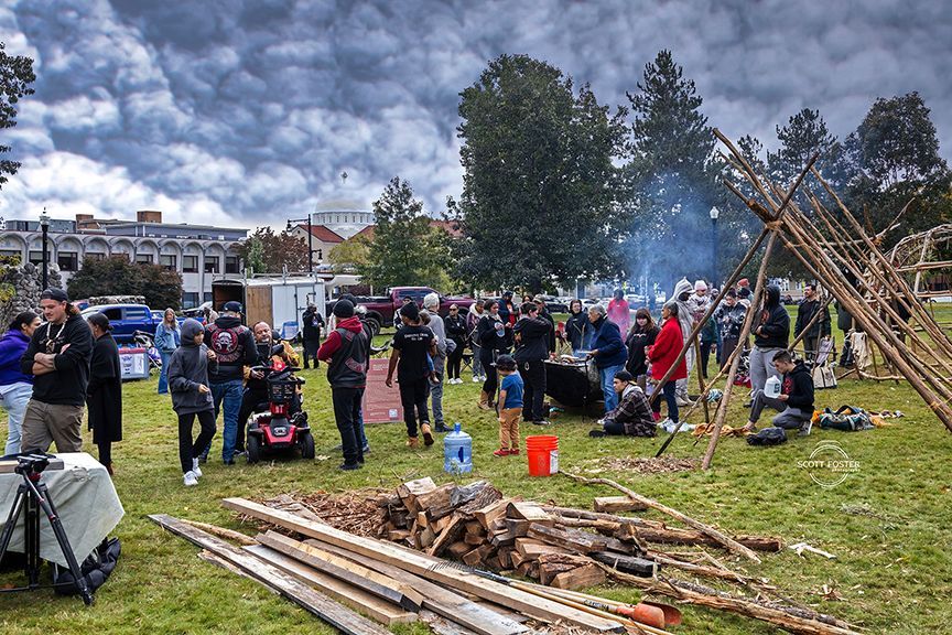 Large outdoor gathering with people near a structure made of poles; a fire burns.