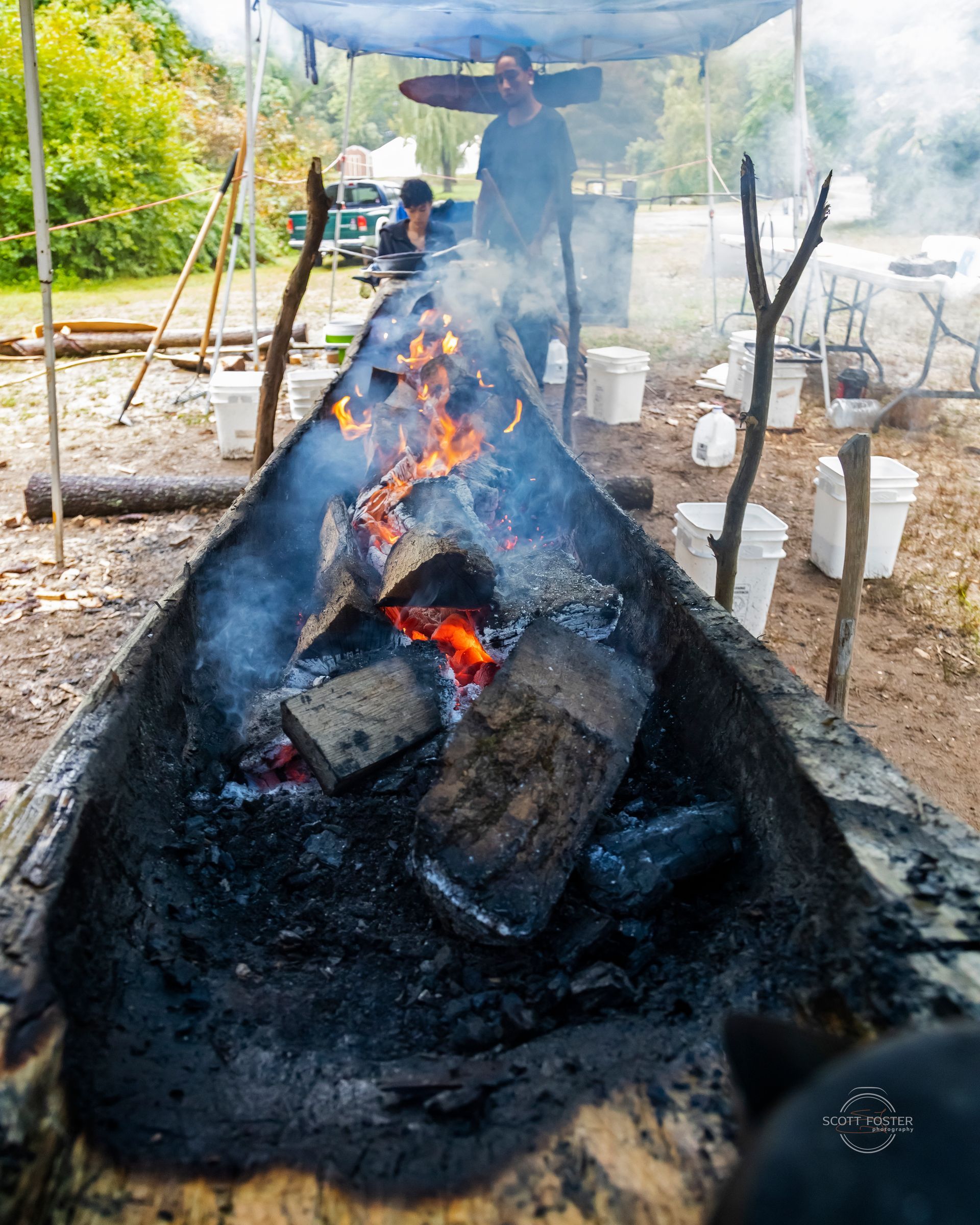Fire burning inside a wooden trough. People are in the background, possibly cooking outdoors.