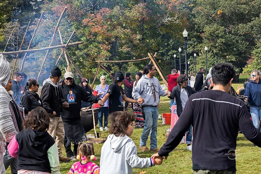 People holding hands, dancing around a fire pit in a park with a wooden structure; sunny day.