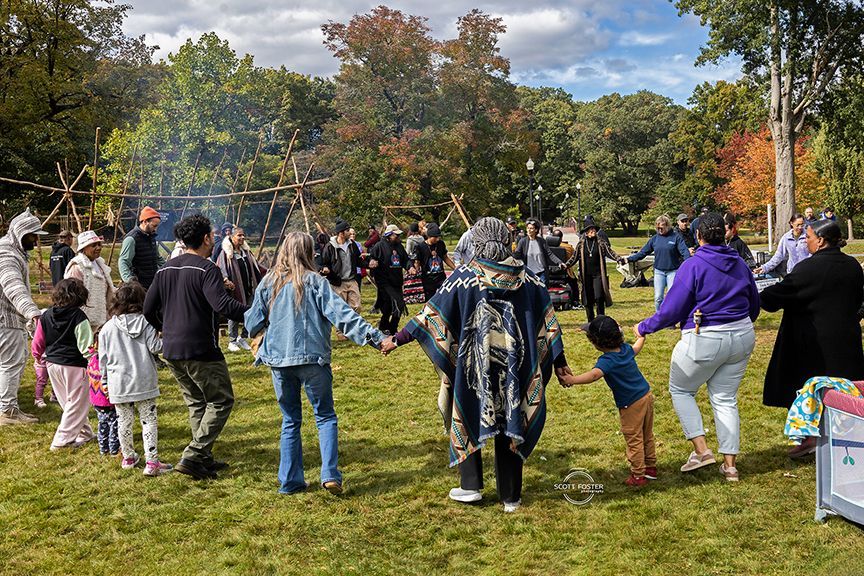 People holding hands in a circle on grass, a ceremonial gathering, with teepee-like structure and trees.