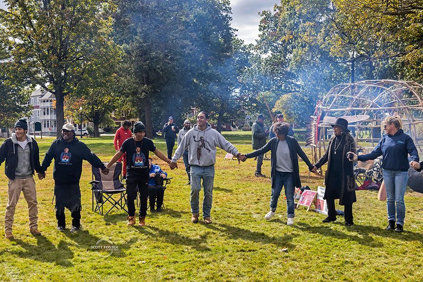 People holding hands in a circle in a grassy park, a fire in the background.