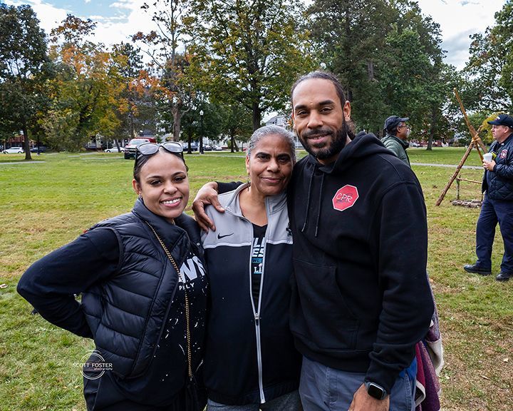 Three people smiling and posing outdoors. Woman in black vest, woman in gray zip-up, and man in black hoodie.