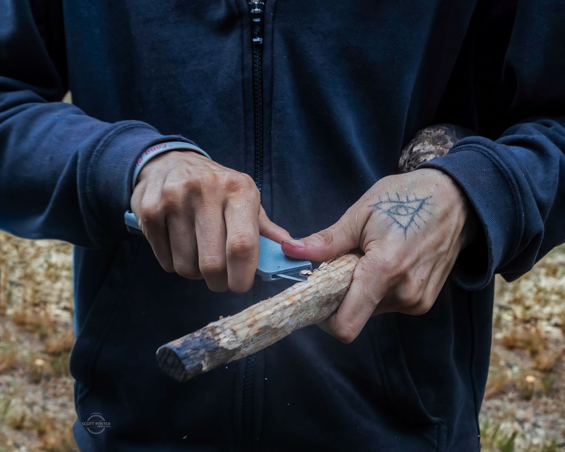 Person carving wood with a knife; hands are visible, wearing a navy jacket and has a hand tattoo, outdoor setting.