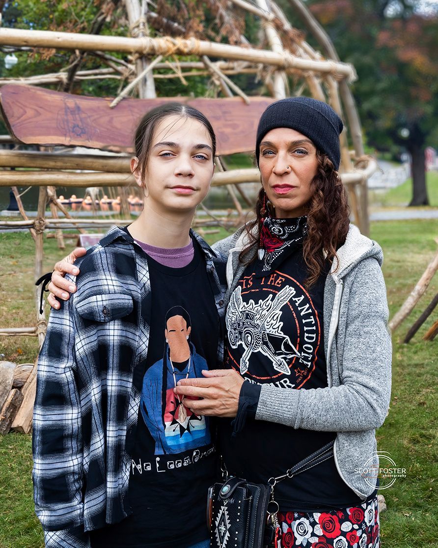 Woman and teen pose outdoors. Teen wears a flannel over a t-shirt. Woman has long hair and a knit hat.
