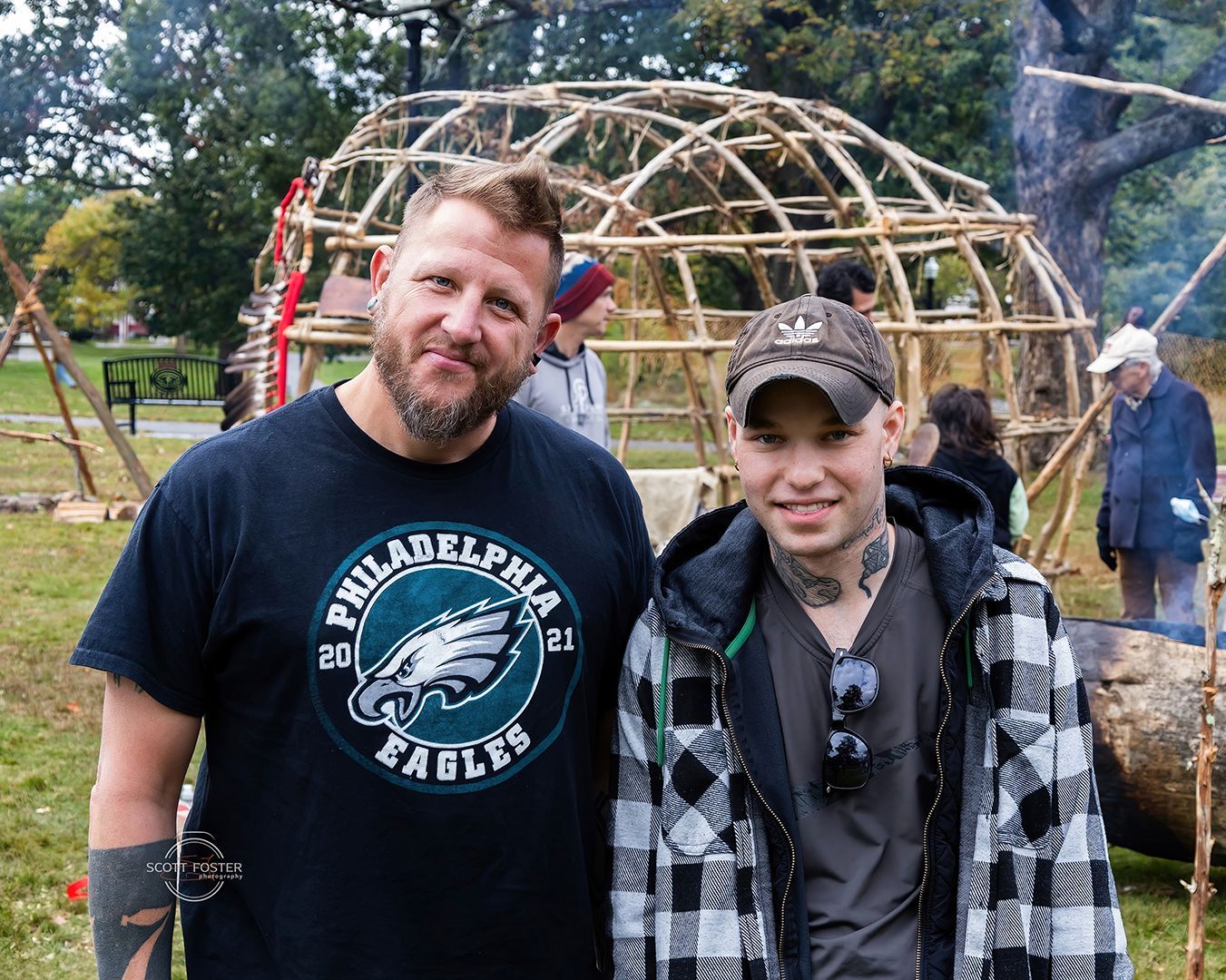 Two men pose outdoors; a wooden dome structure is in the background. One wears an Eagles shirt, the other a plaid jacket.
