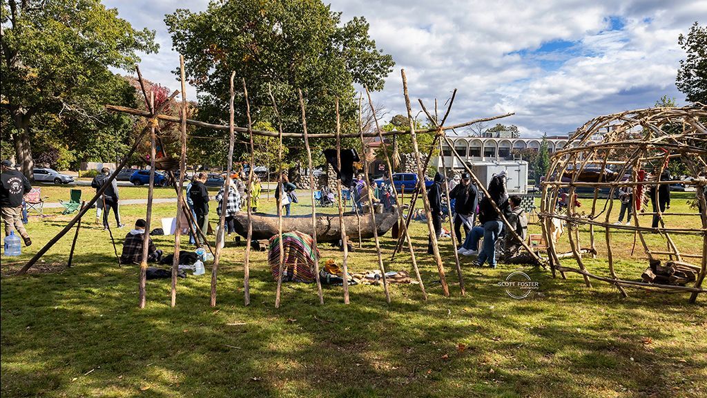 People constructing wooden structures outdoors on a grassy field; trees and cloudy sky in background.