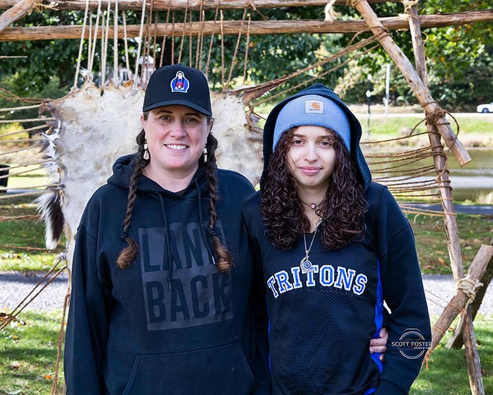 Two people in hoodies pose outdoors near a wooden structure with hides.