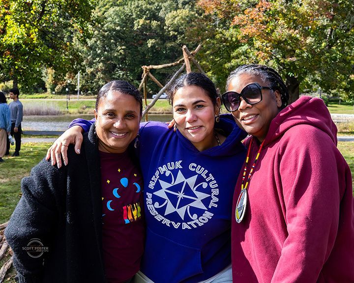 Three people smiling, embracing outdoors. One wears a blue sweatshirt, others red tops. Green trees in background.
