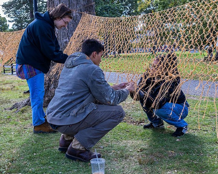 Three people work on a large, woven netting structure outdoors.