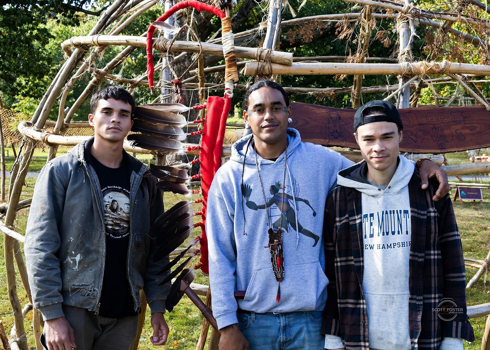 Three people standing in front of a wooden structure decorated with feathers and beads.