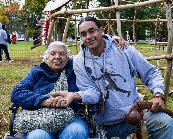Older woman in wheelchair, young man with arm around her, smiling outdoors by a wooden structure.