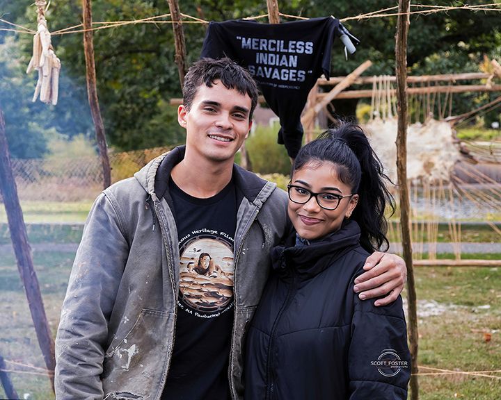 Two people pose outdoors with a wooden structure behind them; one person has an arm around the other.