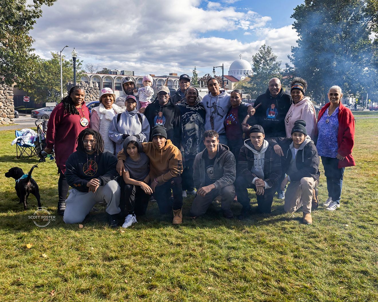 Group of people in a park, posing for a photo. Cloudy sky, trees, and a building are in the background.