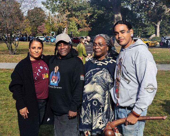 Four people pose outdoors, smiling. One holds a staff. The background is a park with cars and trees.