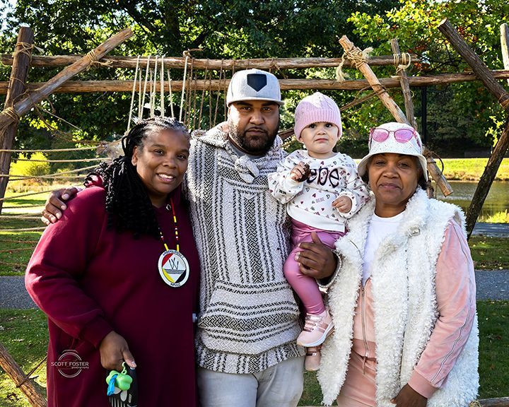 Family posing outdoors near wooden structure. Woman, man, and child smiling; another woman also present.