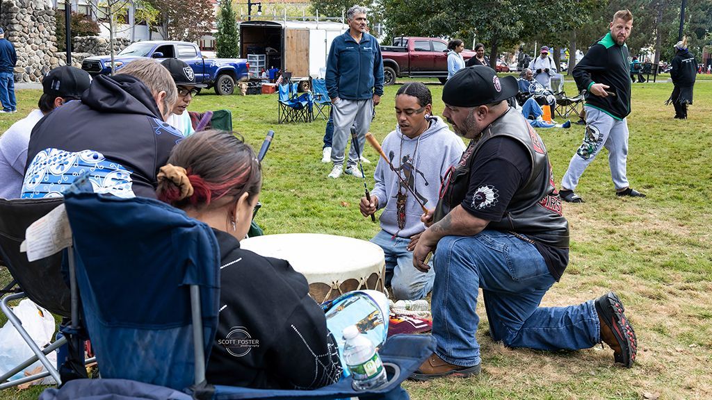 Group of people gathered outdoors, some seated, others standing. A man kneels, holding drumsticks, with others around a drum.