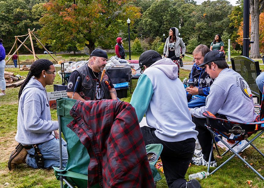 A group of people sitting in a park, talking and looking down. Some are in hoodies; a fire pit is in the background.