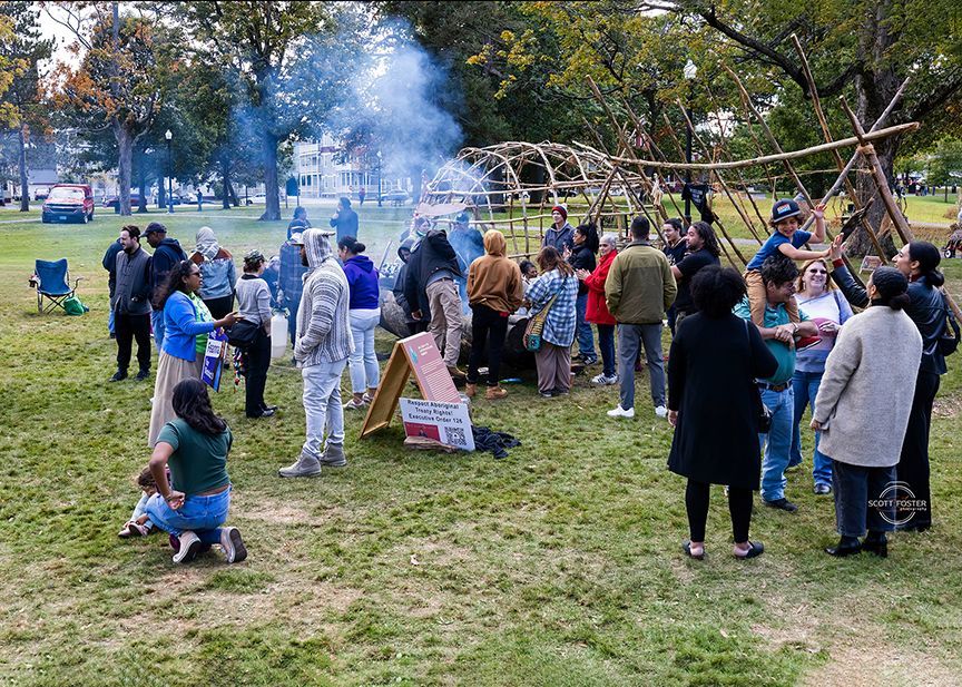 People gather around a fire and wooden structures in a park. Smoke rises.