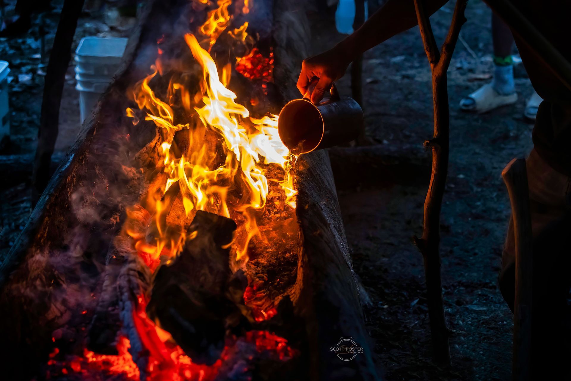 A person pouring liquid from a wooden bowl into a blazing campfire at night.