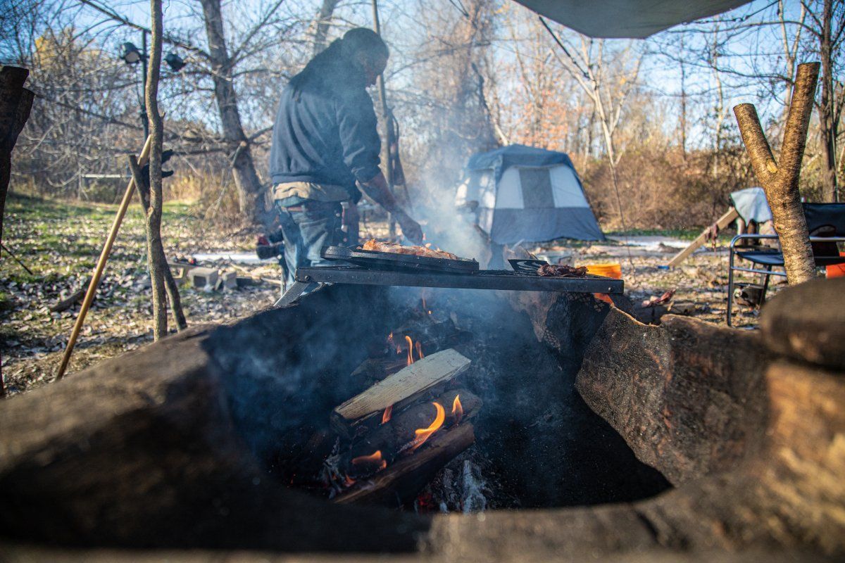 Person cooking over fire in a rustic outdoor setting, smoke rising. Tent in background.