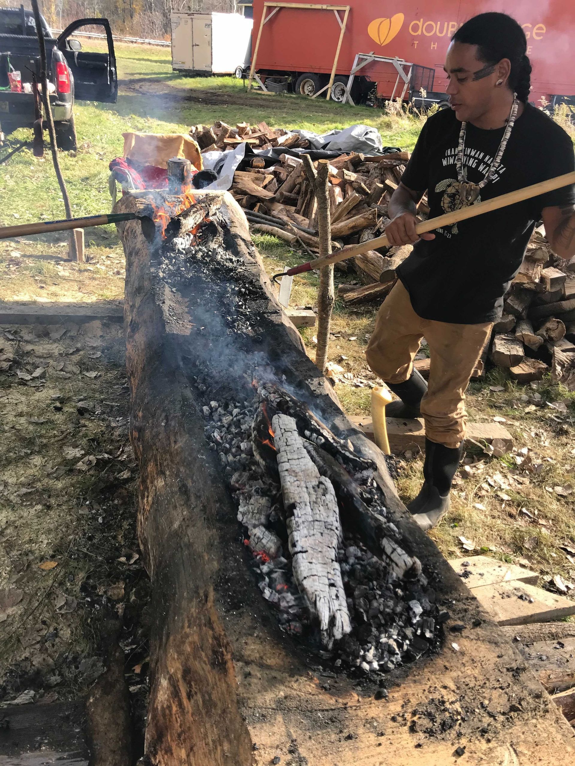 Man tending a fire in a large log, using a long stick. Outdoors, smoky.