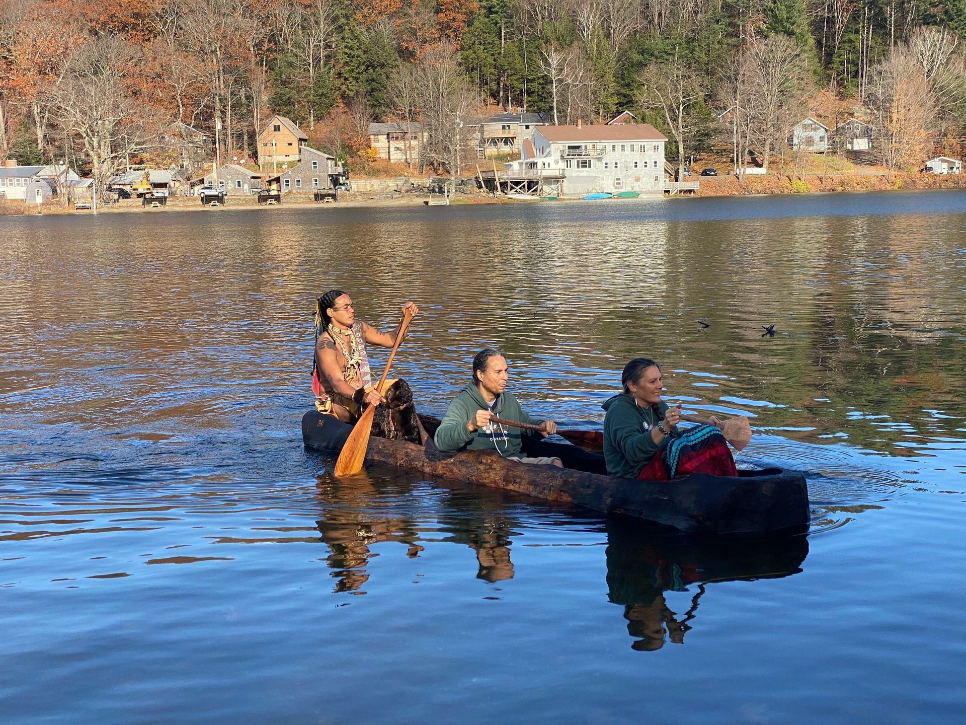 Three people paddle a log canoe on a lake, buildings and trees in the background.