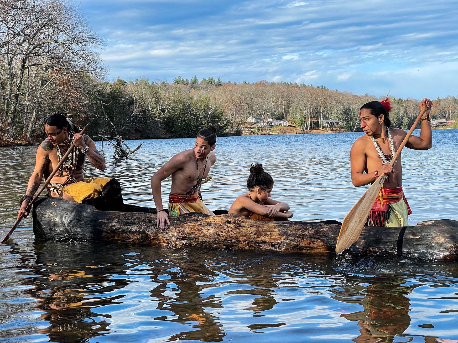 Four people in a wooden canoe on a lake, paddling. One person is resting, two are paddling, and one is steering. Sunny day.