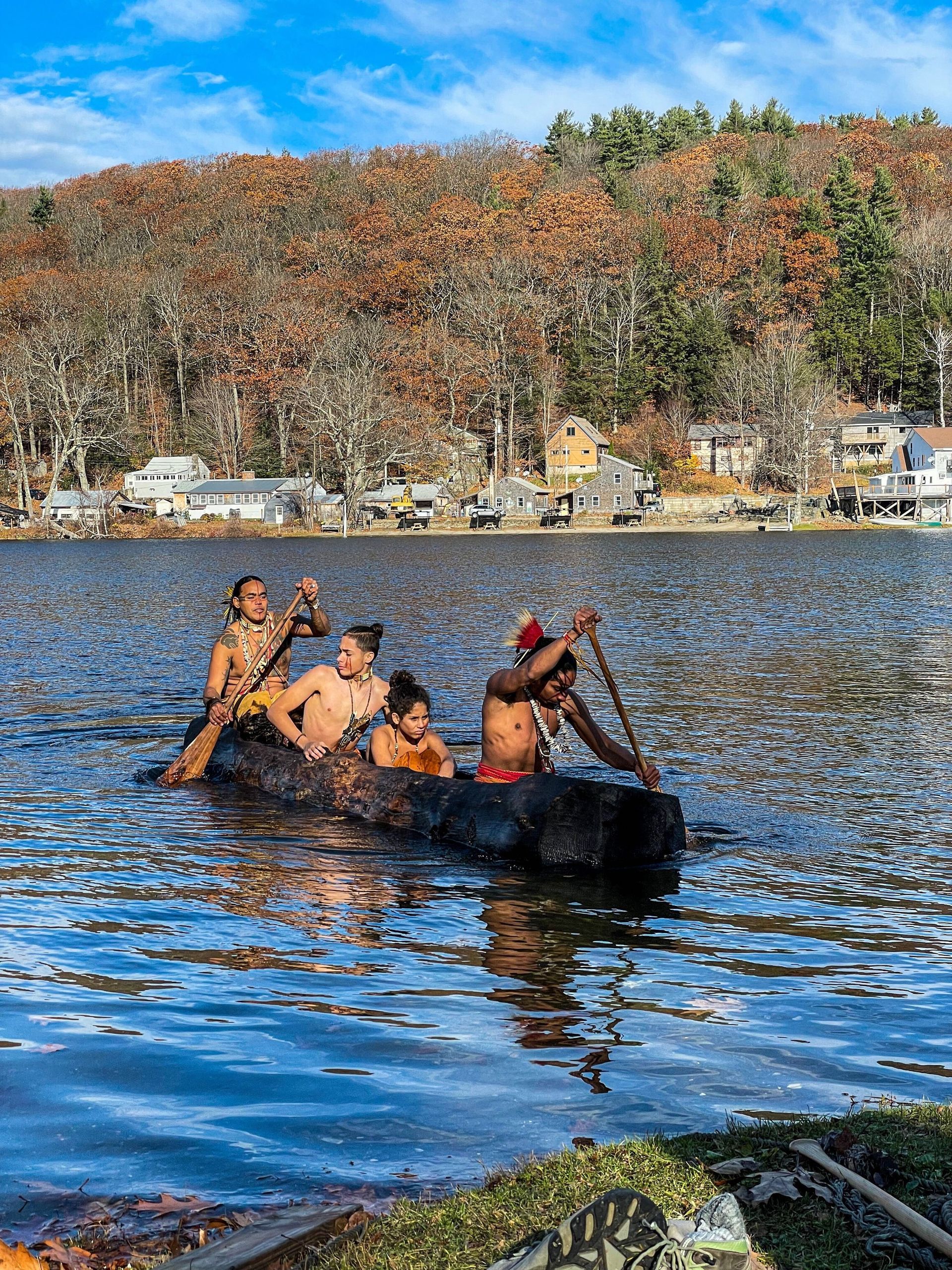 Four people paddling a log canoe on a lake, with a treeline and houses in the background.