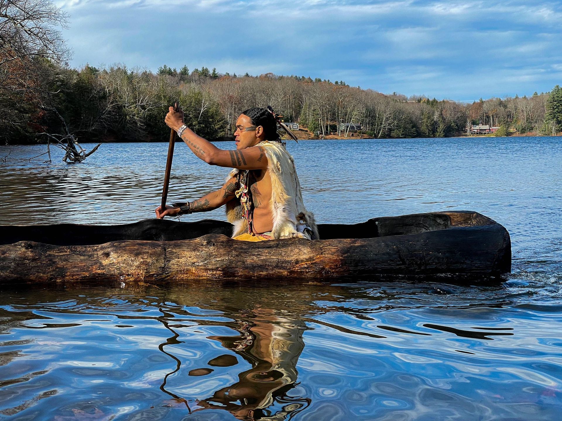 Person paddles a wooden canoe on a lake, holding a paddle. Overcast day with trees in the background.