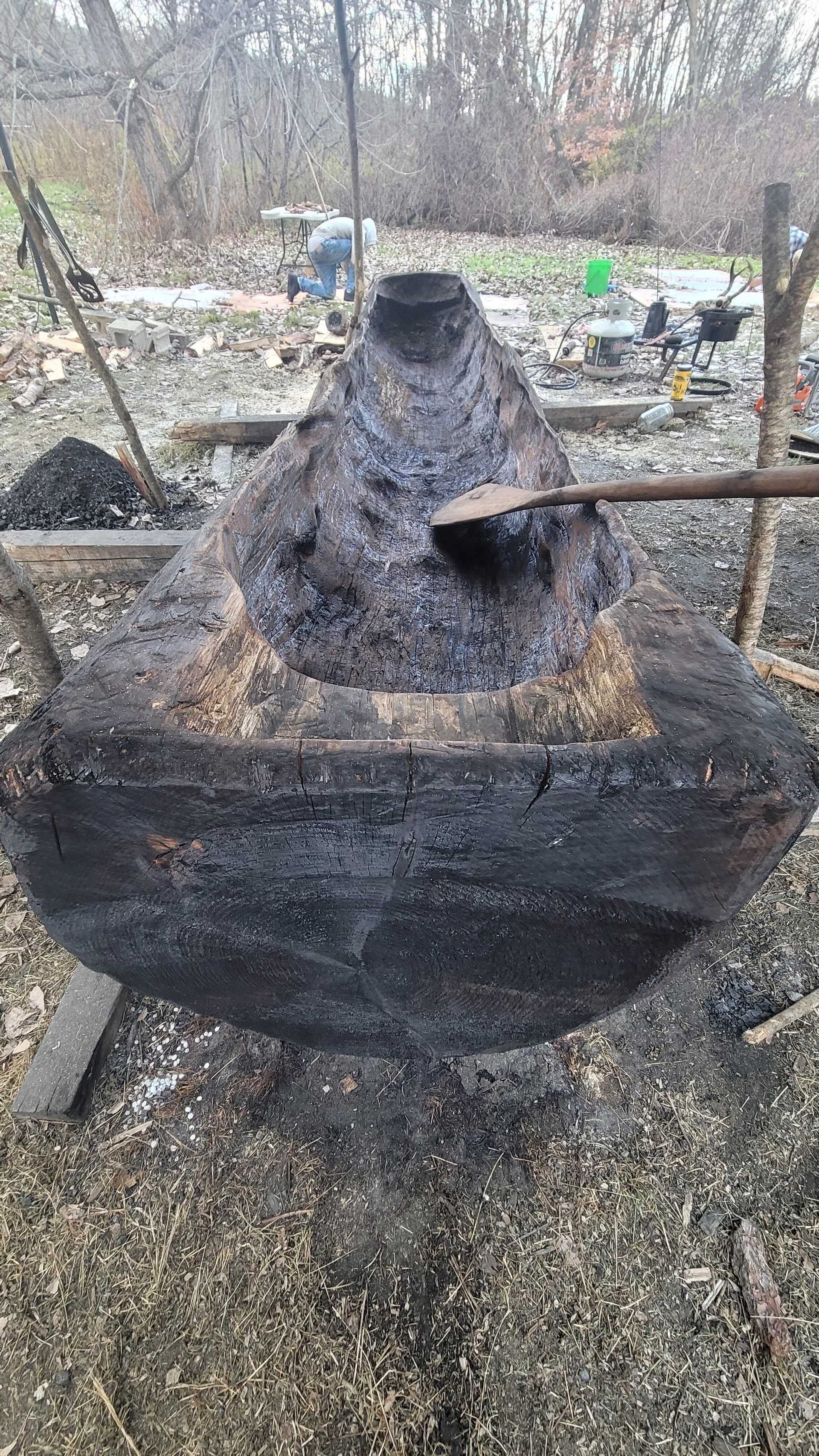 Large, dark, wood-fired container, likely for cooking. A person tends to it with a long-handled tool, outdoor setting.