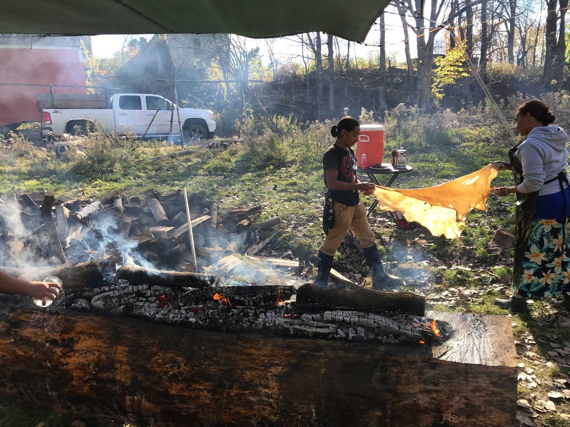 People tending a fire outdoors, a truck and trees visible in the background. One person holds a yellow cloth.
