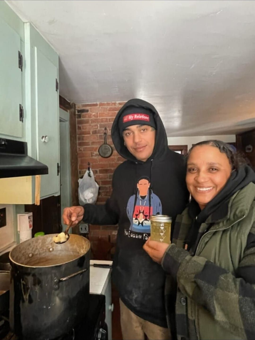 Two people cooking in a kitchen. One stirs a pot, the other holds up a jar of liquid; both smile.