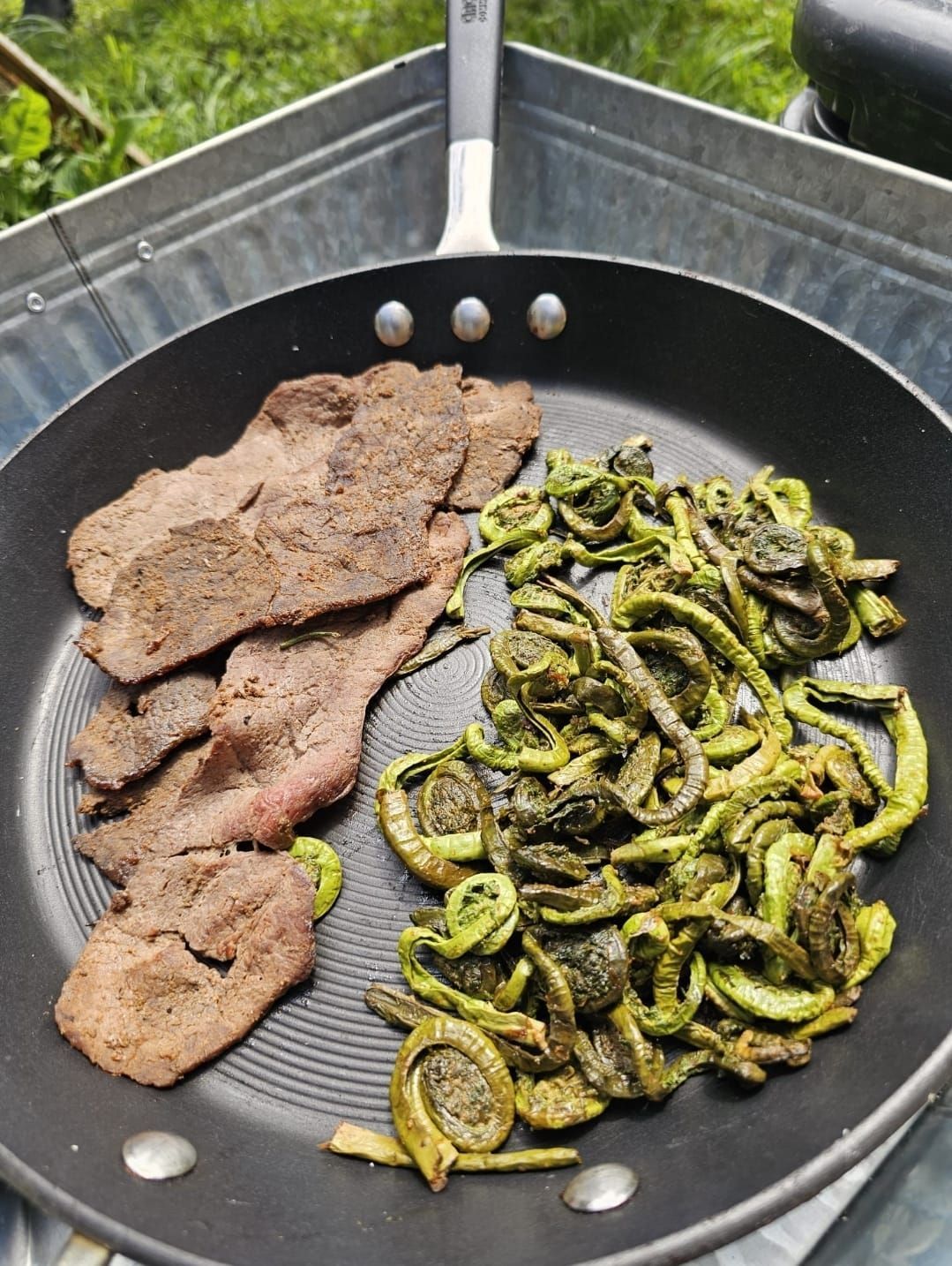 Sautéed meat and green vegetables in a black frying pan outdoors.