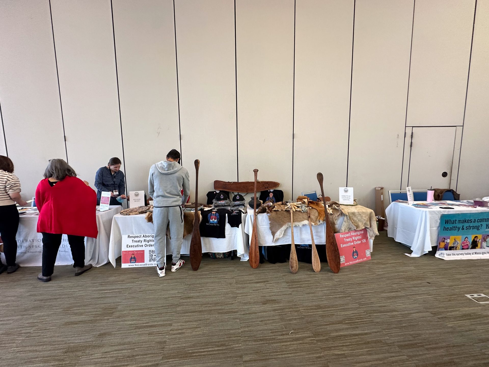 People browsing tables with crafts at an event. Paddles, shirts, and signs are visible.