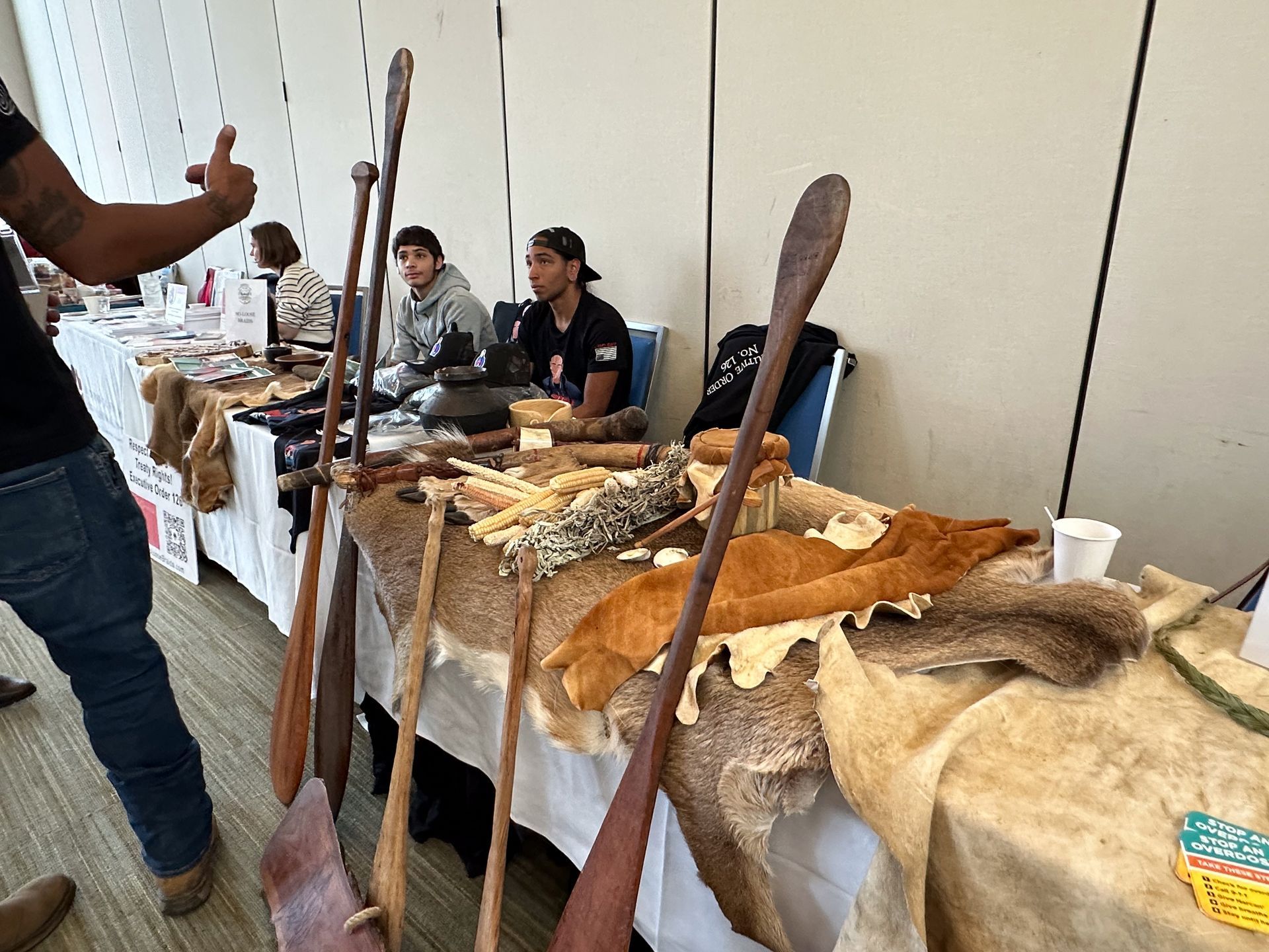 Display table with handcrafted items, including paddles, furs, and people at an event.