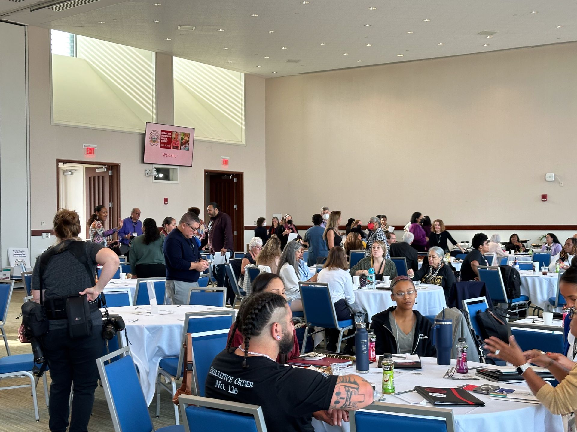 Large room with round tables, people seated and standing, some conversing. White tablecloths, blue chairs.