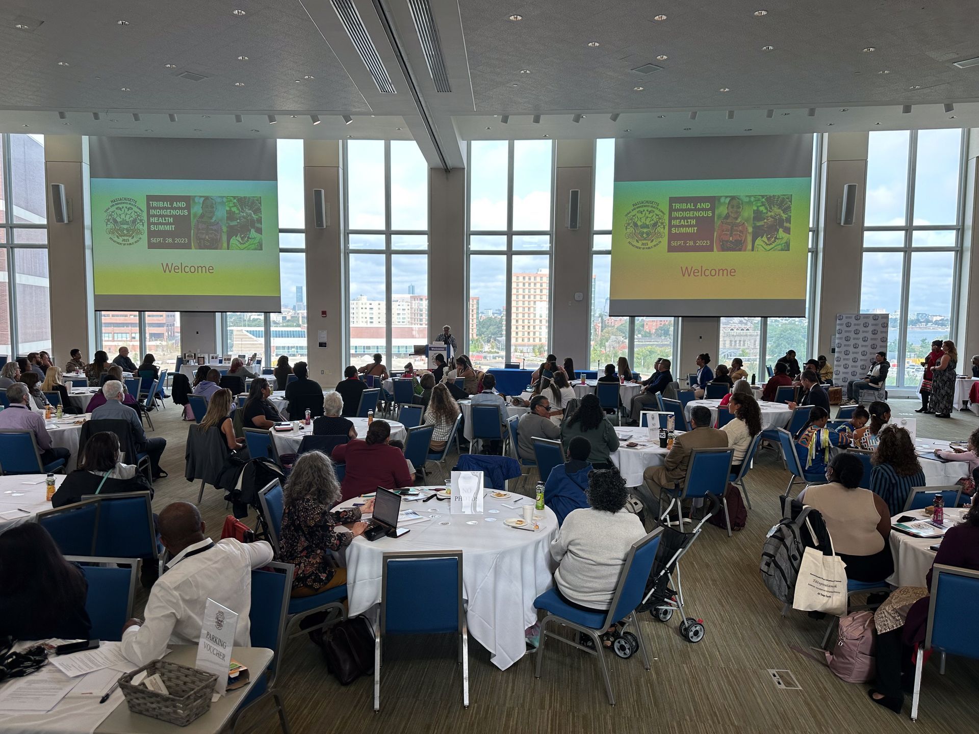 Large conference room with many people at round tables, two projectors displaying images, tall windows.