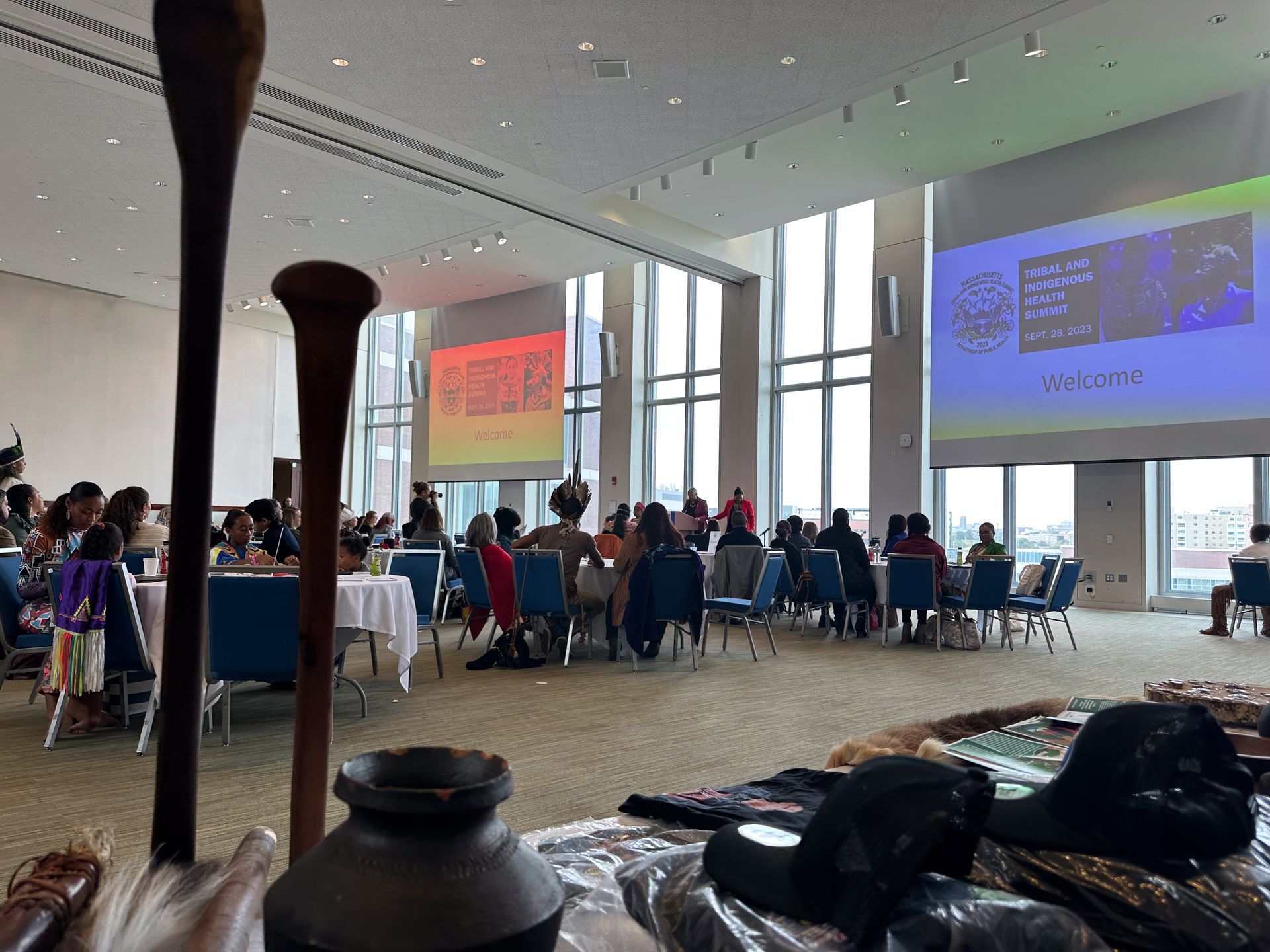Conference hall with attendees, tables, and projections. Foreground: artifacts.