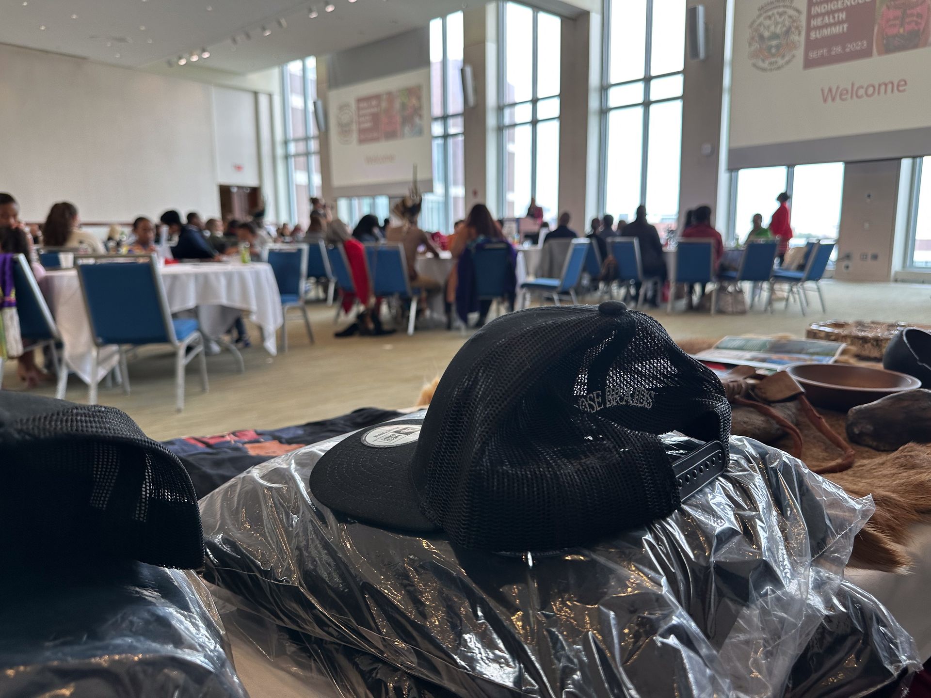 Black hats on a table in front of people seated at tables in a brightly lit room.