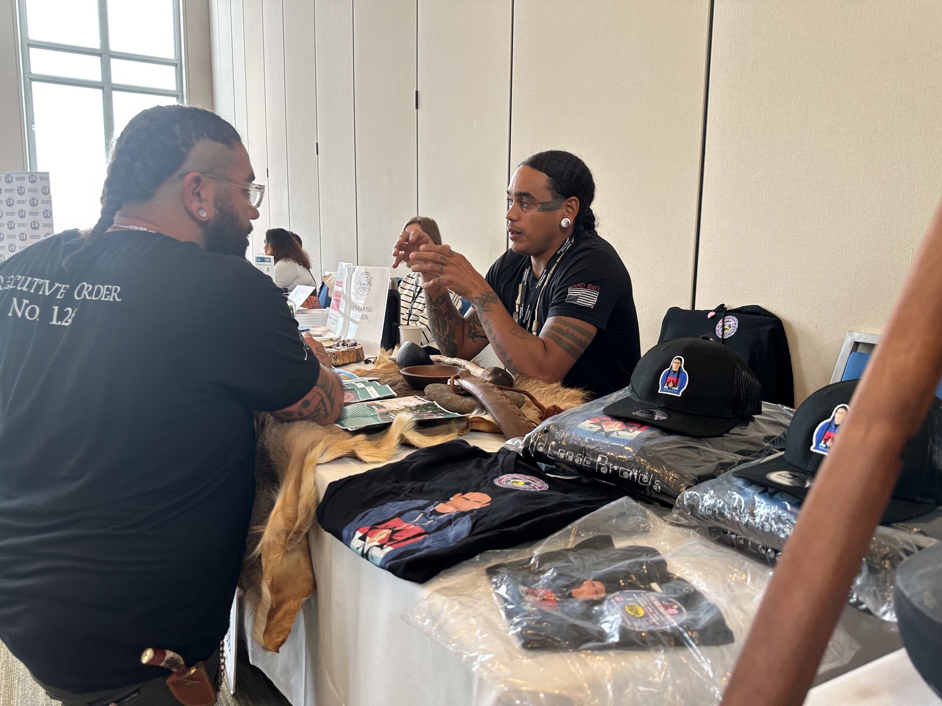 Two men at a vendor table with clothing and hats; one is speaking and gesturing.
