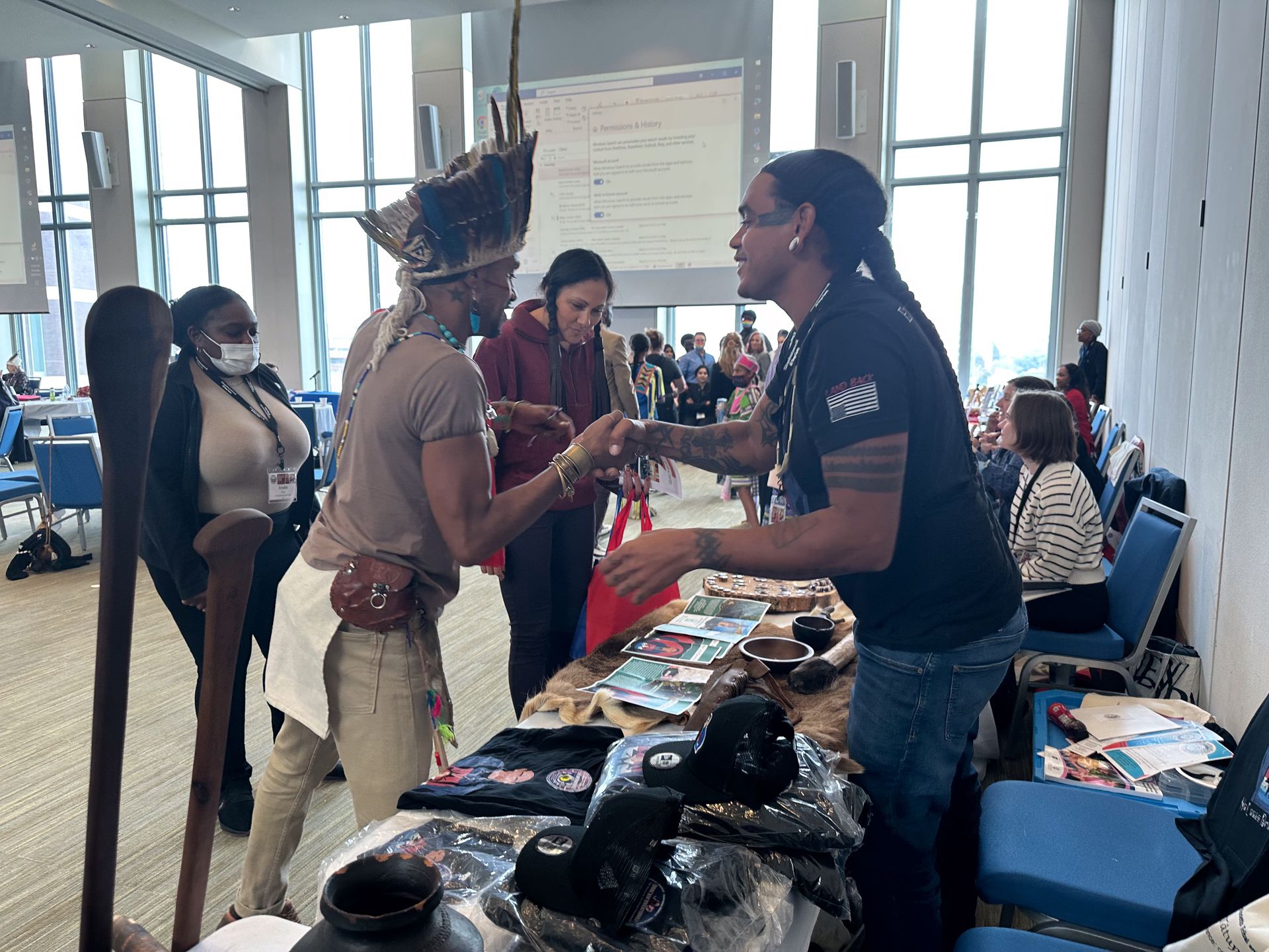 Two men shaking hands at a vendor's table. One wears tribal headwear. Conference setting with tables and chairs.