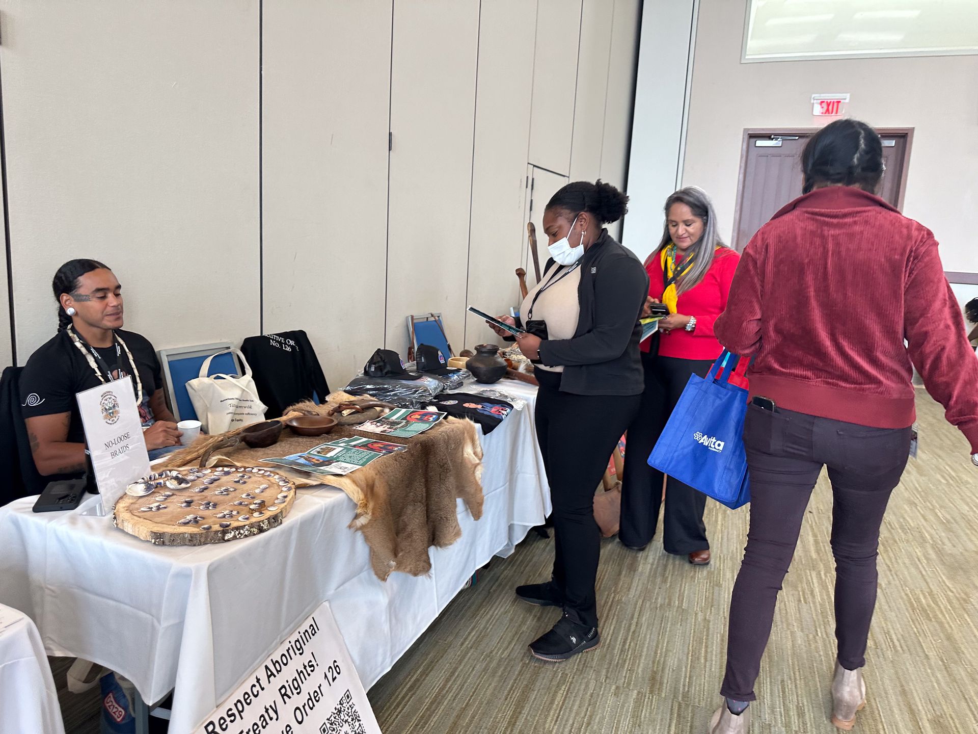 People browsing a vendor's table with crafts. The vendor is seated. A woman in a red jacket walks by.