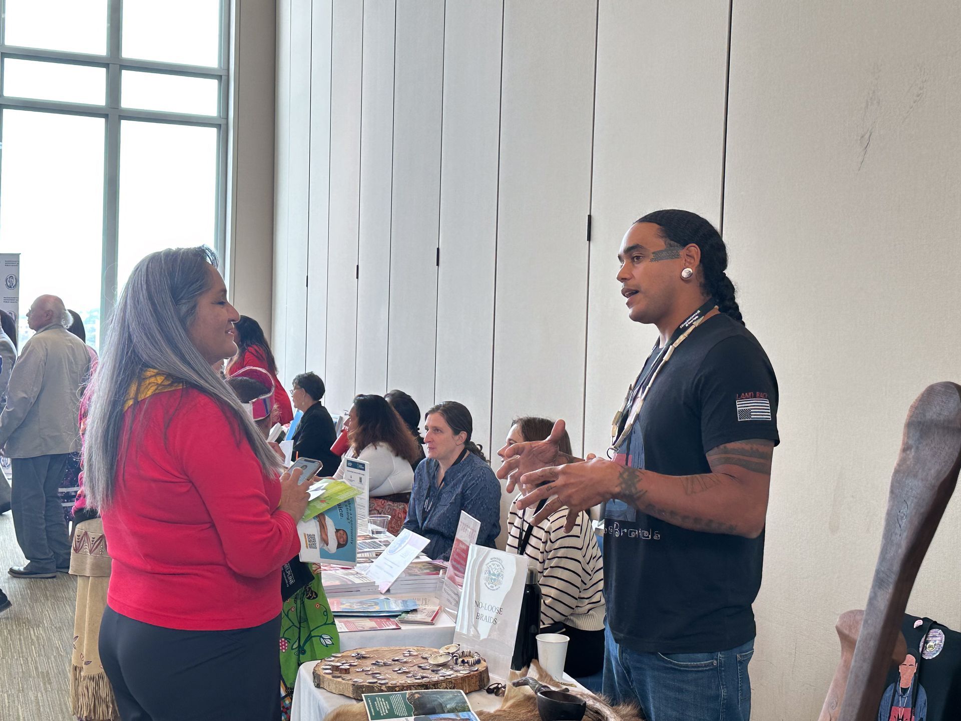 Two people talking at a vendor table. Others nearby, indoors.